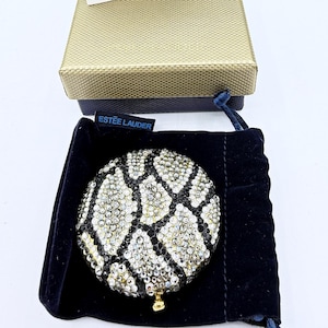 Könnte beinhalten: Ein Estée Lauder Kompaktspiegel mit Schlangenmuster, besetzt mit glitzernden Kristallen. Der Kompaktspiegel befindet sich in einem schwarzen Samtbeutel, mit einer goldfarbenen Schachtel und einer Produktinformationskarte.