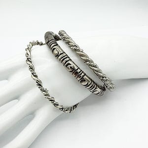 May include: A set of three silver-toned metal bangles. One bangle features a thin twisted rope design, another is a thicker band with embossed circular and linear patterns, and the third is a textured rope-style band with small bead accents.