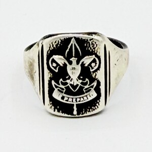 May include: Silver-toned ring with a rectangular face featuring a black emblem. The emblem includes a fleur-de-lis, a dove, and the word "PREPARED". The ring has a slightly worn appearance.