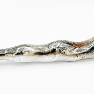 May include: A silver-toned metal handle with an irregular, organic, and undulating shape. The surface features a polished finish with areas of dark tarnish and oxidation. One end has a decorative ribbed band, while the other end has a small metal loop.