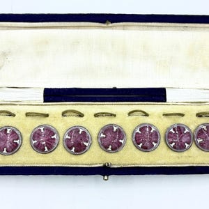 May include: A set of seven antique buttons in a velvet-lined box. The buttons are round with a silver-colored border and a burgundy-colored center with a cross design. The box is navy blue with a cream-colored interior.