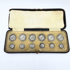May include: A set of antique silver buttons displayed in a black leatherette box with a cream-colored interior. The buttons feature detailed relief designs and are arranged in two rows. The box is open, revealing the buttons.