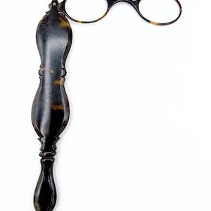 May include: Antique pince-nez eyeglasses with a dark, patterned frame and handle. The glasses feature round lenses and a tortoiseshell pattern on the frame. The handle is long and ornate, with a dark, polished finish. The overall design is vintage and elegant.