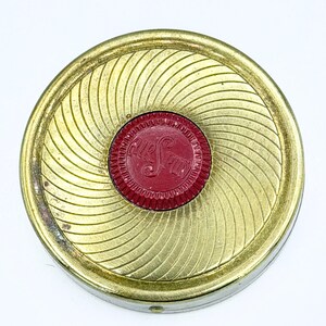 May include: A vintage compact with a gold-toned, spiral-patterned exterior. The center features a red circular emblem with the text "Air Spun". The compact is round and appears to be made of metal.