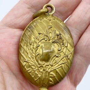 May include: A gold-toned oval locket with a floral design. The locket has a hinged lid and a loop for attaching a chain.