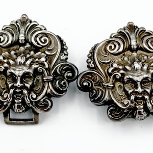 May include: Two antique silver-toned belt buckles. Each buckle features a detailed face with a beard, surrounded by ornate scrollwork and floral designs. One buckle has a rectangular clasp.