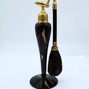 Può includere: Un flacone di profumo in vetro nero con dettagli dorati e un vaporizzatore nero e dorato. Il flacone ha una base rotonda e un collo lungo e sottile. Il flacone è sormontato da un vaporizzatore nero e dorato e da un fiocco nero.