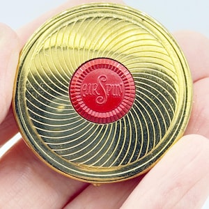 May include: A vintage gold-coloured compact with a red centre. The compact has a spiral design on the lid and the words "Air Spin" are embossed in the centre. The compact is round and appears to be made of metal.