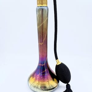 May include: An antique perfume bottle with a bulb atomizer. The glass bottle has a pearlescent finish with gold and purple hues. The atomizer and accents are gold-toned, and the bulb and tassel are black.