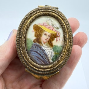 Peut inclure: Compact ovale antique en laiton avec un portrait miniature d'une femme portant un chapeau jaune avec un ruban et des plumes roses. Le portrait est placé sur un fond vert et blanc. Le compact est tenu dans une main.