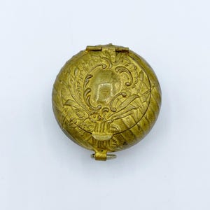 Puede incluir: Una pequeña caja redonda de metal de color dorado con un diseño floral ornamentado en la tapa. La caja tiene una tapa con bisagras y un pequeño lazo en el lateral.