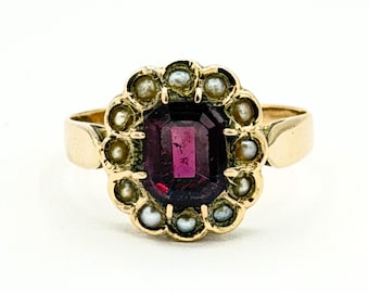 May include: A gold ring featuring a large, rectangular, deep purple gemstone at the center, surrounded by a halo of small pearls. The ring's band is a simple, polished gold. The ring is set against a white background.