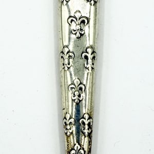 May include: A silver-toned metal handle featuring a repeating embossed fleur-de-lis pattern. The elongated, tapered handle has a polished finish with visible dark oxidation in the recessed areas of the metal design, creating a high-contrast look.