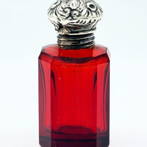 May include: A small, antique red glass bottle with a silver-colored, ornate stopper. The bottle has a rectangular shape with beveled sides. The stopper is highly detailed with a floral design. The bottle is empty and sits against a white background.