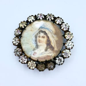 May include: A vintage brooch with a painted portrait of a woman in a white dress and a blue hat. The portrait is surrounded by a circle of clear rhinestones.