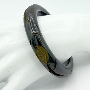 May include: A dark charcoal gray bangle bracelet with a smooth, polished finish. The surface features carved, recessed leaf-like patterns that reveal a lighter, brownish-gold tone within the grooves, creating a subtle contrast in color and texture.