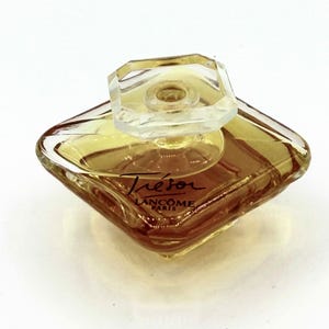 Puede incluir: Un frasco de perfume de vidrio en forma de diamante lleno de líquido de color ámbar. El frasco cuenta con un tapón de vidrio transparente facetado y el texto Trésor LANCOME PARIS impreso en negro en la parte frontal del recipiente.
