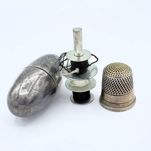 Op de afbeelding: Een verzameling naaigerei op een witte achtergrond. De set bevat een zilveren vingerhoed, een zilveren draadhouder, een zilveren ovale naaldenkoker en spoelen met zwart en wit garen.