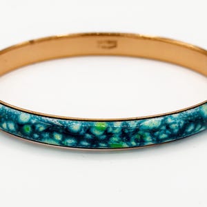 May include: A copper-coloured bangle bracelet with a turquoise and green speckled enamel design. The bracelet has a smooth, polished finish. The jewellery is a classic accessory. The diameter is approximately 6.5 cm.