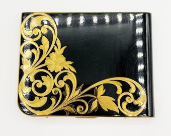 May include: A rectangular black case with a glossy finish, featuring an intricate gold-colored floral and scrollwork design that extends from the left side across the bottom edge, accented by a thin gold-colored metal border.