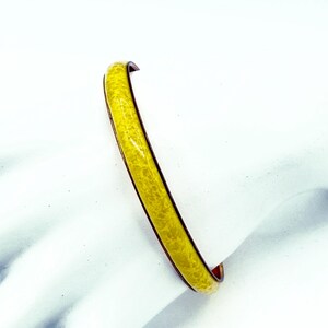 May include: A yellow bangle bracelet with a speckled pattern. The bracelet has a dark brown interior and a smooth, curved design. The yellow colour is bright, making it a stylish accessory.
