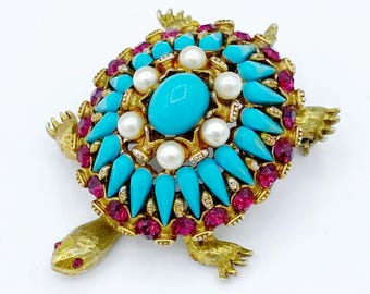 May include: A gold tone turtle brooch featuring a turquoise and red rhinestone design. The turtle's shell is adorned with a large turquoise cabochon surrounded by smaller turquoise stones and red rhinestones. The turtle's legs and head are detailed with gold tone accents.