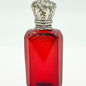 Puede incluir: Un frasco de perfume de vidrio rojo rubí con un tapón plateado y ornamentado. El frasco tiene forma rectangular con bordes biselados. El tapón presenta un diseño floral detallado. El frasco está vacío y sobre un fondo blanco.