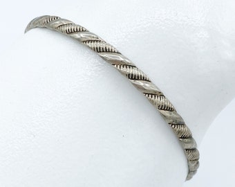 Könnte beinhalten: Ein silberfarbenes Armband mit einem gedrehten Seildesign. Das Armband hat eine strukturierte Oberfläche und wird vor einem weißen Hintergrund gezeigt. Das Schmuckstück ist ein schlichtes, elegantes Accessoire.
