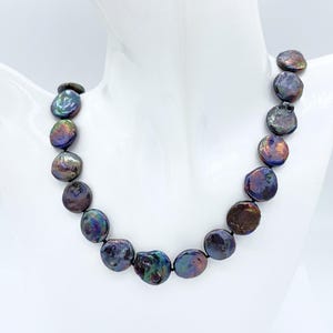 May include: A necklace featuring iridescent, coin-shaped pearls. The pearls display a range of colors, including purple, blue, and green, strung together on a dark cord. The necklace is displayed on a white mannequin.