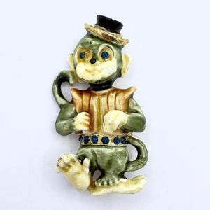 以下が含まれることがあります： 黒いシルクハットと金の帯が付いたヴィンテージモンキーブローチ。 猿は緑色で、顔と手はクリーム色です。 青いラインストーンの目と金と茶色のハーモニカが付いています。 青いラインストーンのベルトがウエストを囲んでいます。