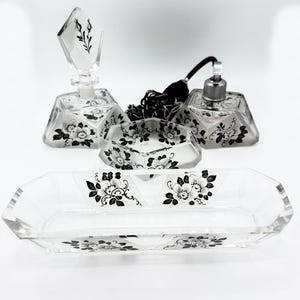 Pode incluir: Um conjunto de artigos de higiene pessoal de vidro transparente vintage, incluindo dois frascos de perfume, um prato pequeno e uma bandeja longa. Cada peça é decorada com desenhos florais pretos. Os frascos de perfume têm formas geométricas e rolhas decorativas.