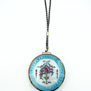 May include: Round, vintage pendant with a floral design. The pendant features a light blue border and a central image of a vase filled with pink and purple flowers. It has a gold-colored metal frame and a braided cord.