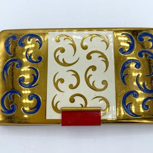 Op de afbeelding: Een gouden compact met een wit centraal paneel en blauwe en gouden draaiende ontwerpen. De compact heeft een rode sluiting.