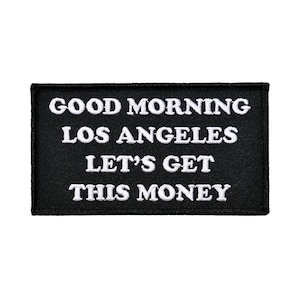 Op de afbeelding: Een rechthoekige zwarte patch met witte geborduurde tekst. De tekst luidt: "GOOD MORNING LOS ANGELES LET'S GET THIS MONEY". De patch is waarschijnlijk bedoeld om op kleding of accessoires te worden genaaid of gestreken.