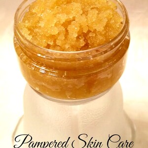 May include: A glass jar filled with a golden brown sugar scrub. The jar is sitting on a white surface with the text "Pampered Skin Care" written in black cursive.