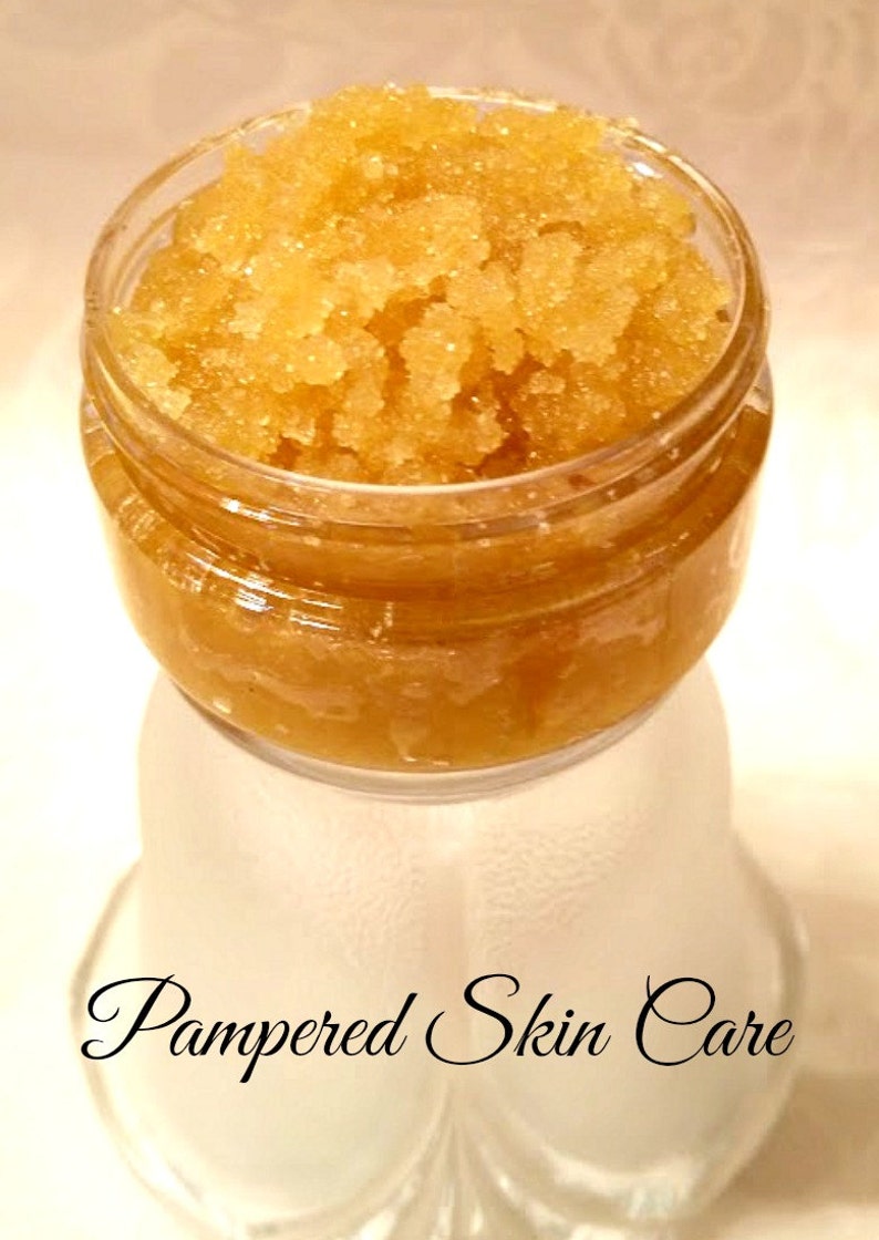 May include: A glass jar filled with a golden brown sugar scrub. The jar is sitting on a white surface with the text "Pampered Skin Care" written in black cursive.
