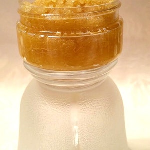 May include: A glass jar filled with a golden brown sugar scrub. The jar is sitting on a clear glass base with the words "Pampered Skin Care" written in black cursive.