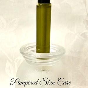 Pu&ograve; includere: Un flacone di vetro trasparente con un flacone roll-on nero e verde all'interno. Il flacone &egrave; appoggiato su una base di vetro bianco smerigliato con le parole "Pampered Skin Care" scritte su di essa.