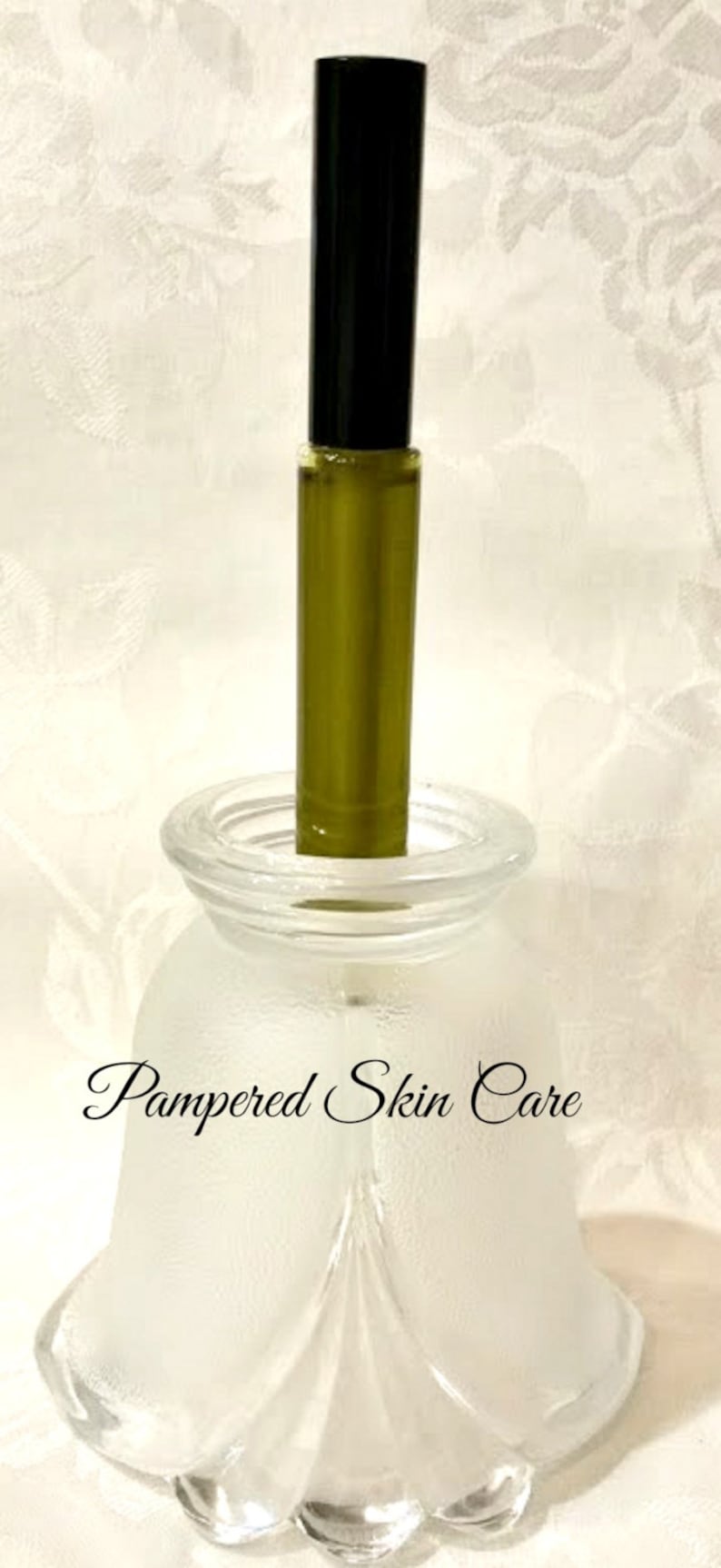 Pu&ograve; includere: Un flacone di vetro trasparente con un flacone roll-on nero e verde all'interno. Il flacone &egrave; appoggiato su una base di vetro bianco smerigliato con le parole "Pampered Skin Care" scritte su di essa.