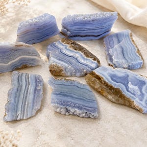 May include: A collection of seven blue lace agate stone slices featuring distinct horizontal banding in shades of light blue, white, and grey. Each piece displays a polished, smooth surface with natural, rough, crystalline edges along the sides.