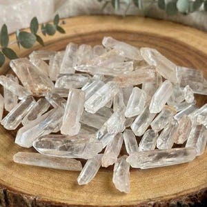 Puede incluir: Un conjunto de puntas de cristal de cuarzo transparente dispuestas sobre una bandeja redonda de madera. Los cristales, de aspecto natural y en bruto, varían en tamaño y forma. La bandeja está rodeada de tela de tonos neutros y vegetación.