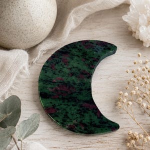 May include: A crescent moon-shaped carving made of polished zoisite stone. The surface features a deep forest green base color with prominent, irregular patches of dark magenta and black mineral inclusions throughout the smooth, flat stone piece.