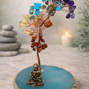 May include: A decorative tree sculpture featuring twisted copper wire branches adorned with multi-colored gemstone chips in shades of yellow, orange, red, green, blue, and purple, set on a round blue agate slice base with a small bronze Buddha.