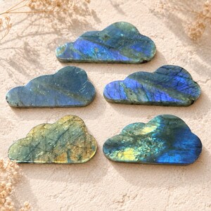 Puede incluir: Cinco tallas planas con forma de nube hechas de piedra labradorita. Cada pieza muestra variaciones minerales naturales con destellos iridiscentes de azul, verde y dorado en las superficies pulidas, resaltando la chatoyance de la piedra.
