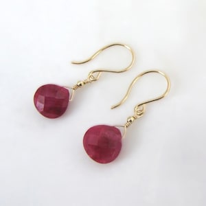 May include: A pair of drop earrings featuring faceted, teardrop-shaped deep pink gemstones suspended from thin gold-toned wire hooks. Each gemstone is attached to the hook with a small gold-toned metal bead and a delicate wire-wrapped loop.