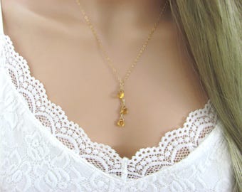 May include: A delicate gold chain necklace featuring three small, faceted, yellow-orange teardrop-shaped gemstones suspended in a vertical cluster. The necklace rests against a white lace-trimmed garment, showcasing a simple and refined design.