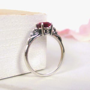May include: A silver ring with a large, oval-cut, red gemstone. The ring has an ornate, filigree design around the gemstone setting. The band is smooth and polished. The ring is displayed on a white surface.