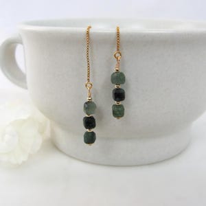 May include: A pair of dangle earrings with a gold chain and three dark green and black square beads. The earrings are displayed in front of a white teacup. A white flower is in the background.