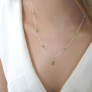 May include: A delicate silver necklace featuring small, round, light green gemstones. The necklace has a simple chain design with several gemstones spaced along the chain and a single gemstone pendant. The necklace is worn against a white background.