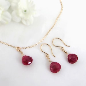 May include: A gold chain necklace and a pair of matching drop earrings, each featuring a single faceted, teardrop-shaped deep pink gemstone pendant. The jewelry set is crafted with delicate gold wire and thin gold chain links.